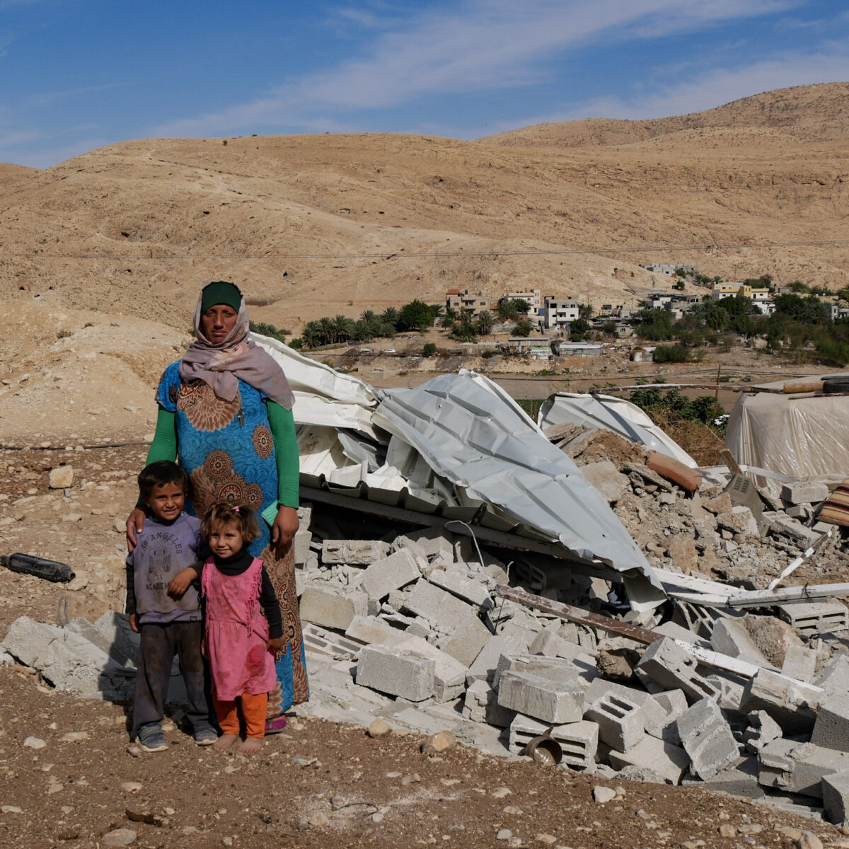 a Palestinian woman stands with her two children in front of their demolished home