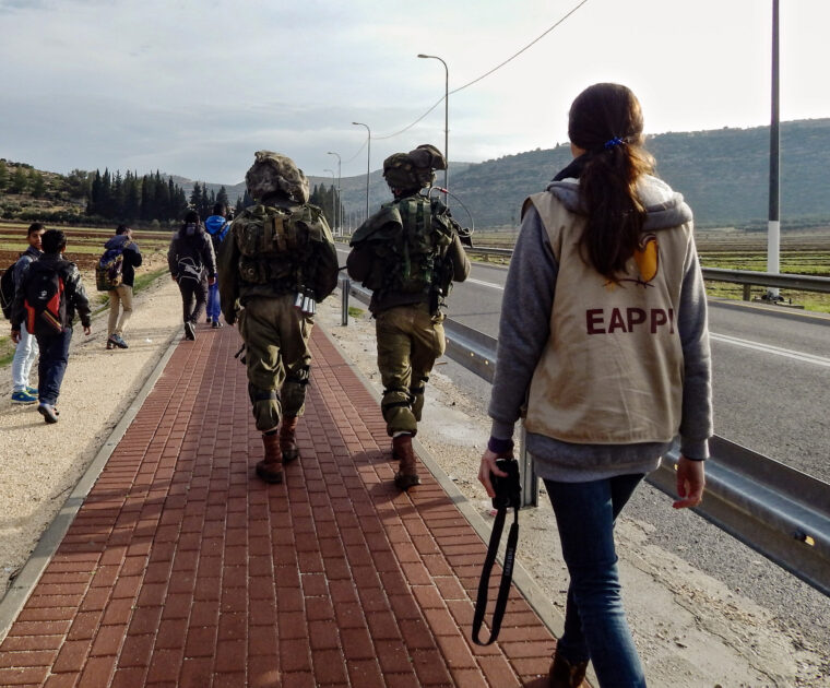 EAs walks with Palestinian children as they go to school past soldiers