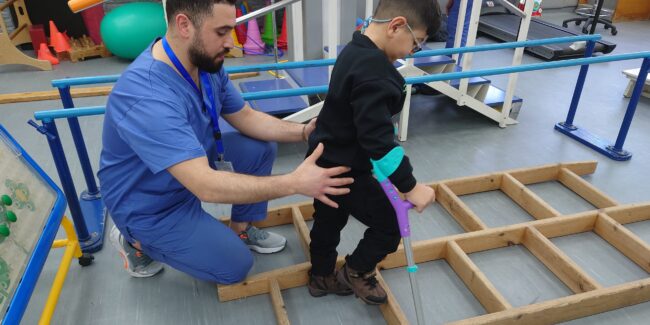 A child with crutches walking across a ladder placed on the floor with the help of a physiotherapist holding his back. The physiotherapist is a man dressed in blue scrubs. They are in a room with lots of physical exercise equipment.