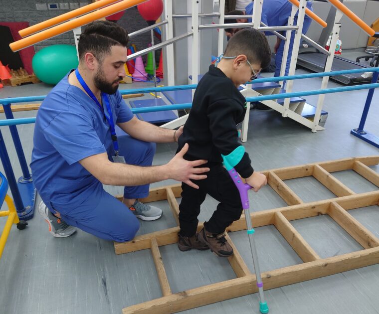 A child with crutches walking across a ladder placed on the floor with the help of a physiotherapist holding his back. The physiotherapist is a man dressed in blue scrubs. They are in a room with lots of physical exercise equipment.