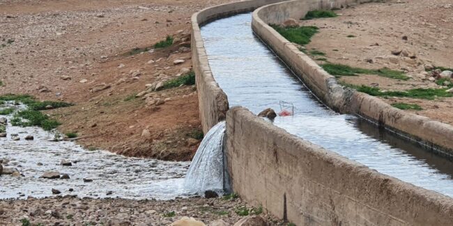 An enclosed water canal running through a desert like land has been broken and water is flowing out of it onto the rocky ground.