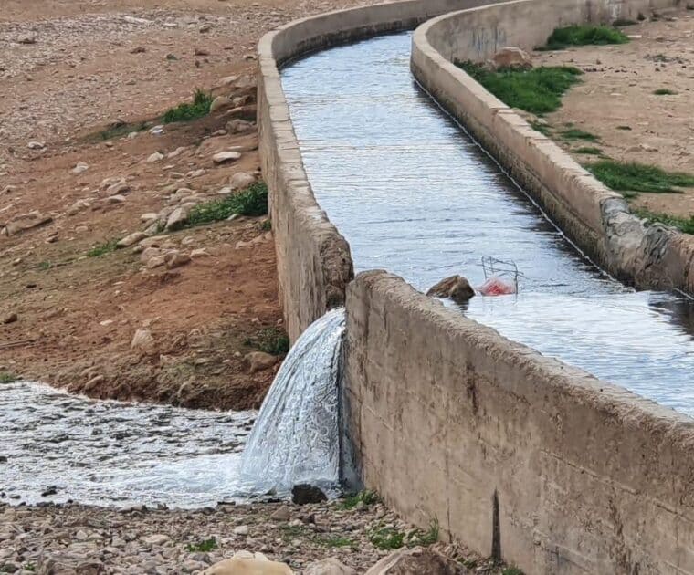 An enclosed water canal running through a desert like land has been broken and water is flowing out of it onto the rocky ground.
