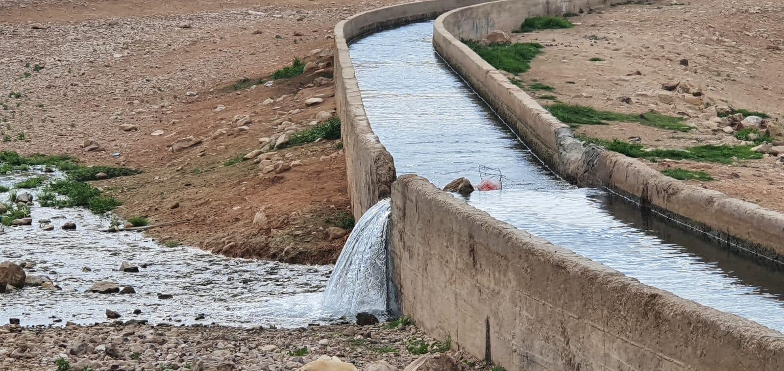 Water blog photo 5 Canalpic-min An enclosed water canal running through a desert like land has been broken and water is flowing out of it onto the rocky ground.