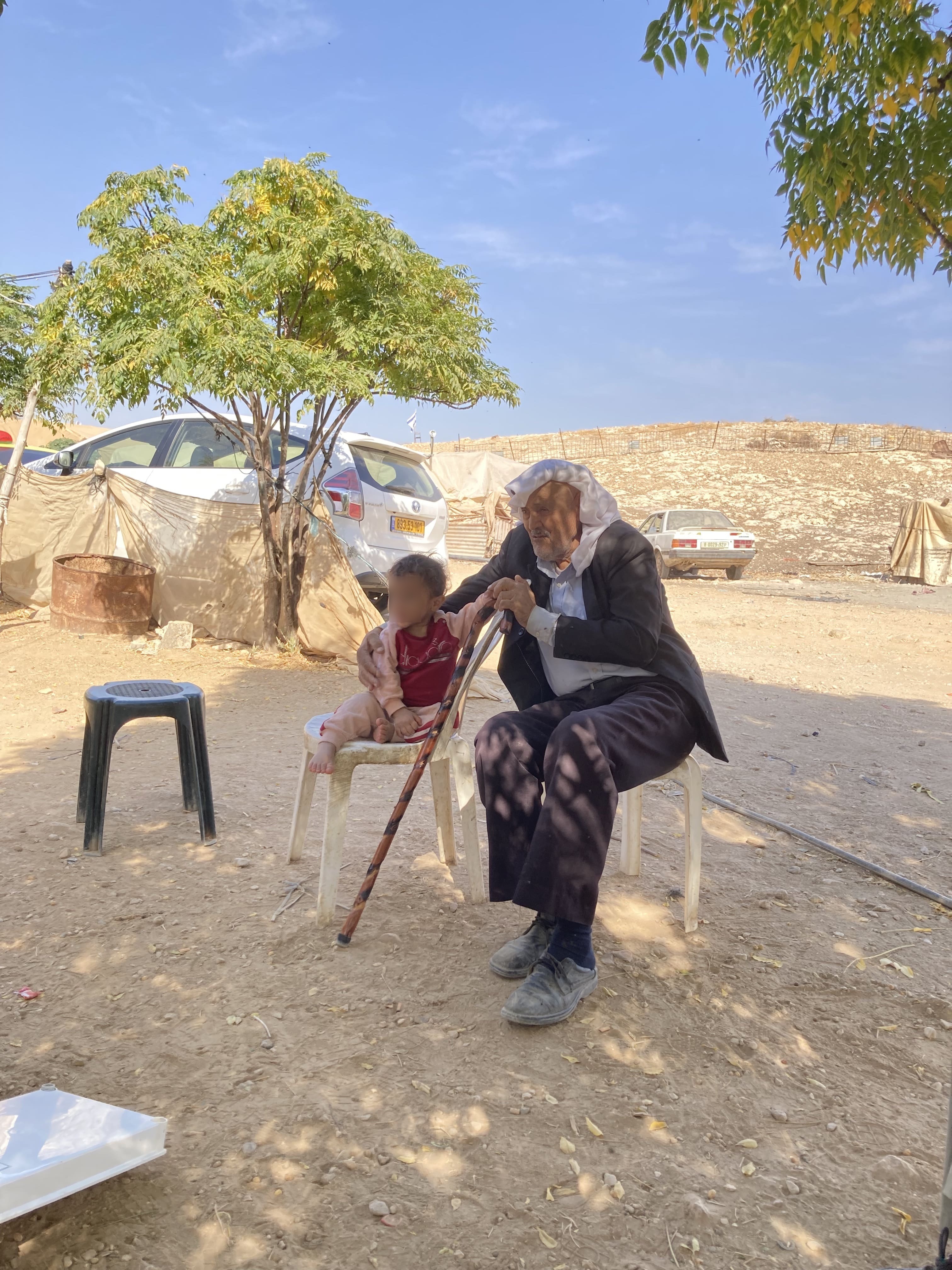 Waterblog photo 6 grandadflag-min (1) (2) An old man sits on a plastic chair with his arm around a small boy, also on a plastic chair. The man is wearing a black suit with a white headwrap. The boy is wearing a light pink matching tracksuit and zip hoodie outfit and a red t-shirt. They are sitting on a dirt patch with three tress and two cars in the background. It's a sunny day, the sky is blue. There is a small hill in the background.