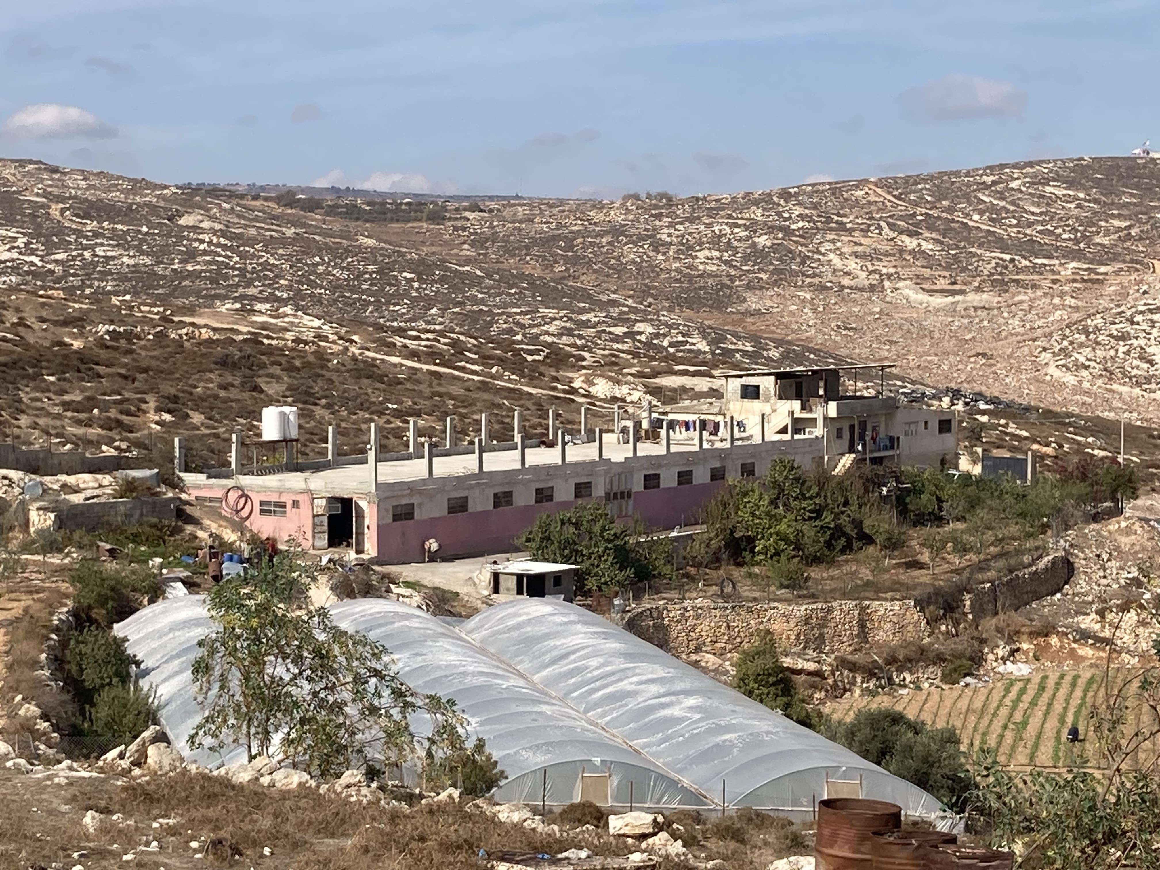 2. The outbuilding where the sheep are kept_EAPPI-min Photo of building in a valley with small window next to three long greenhouses.