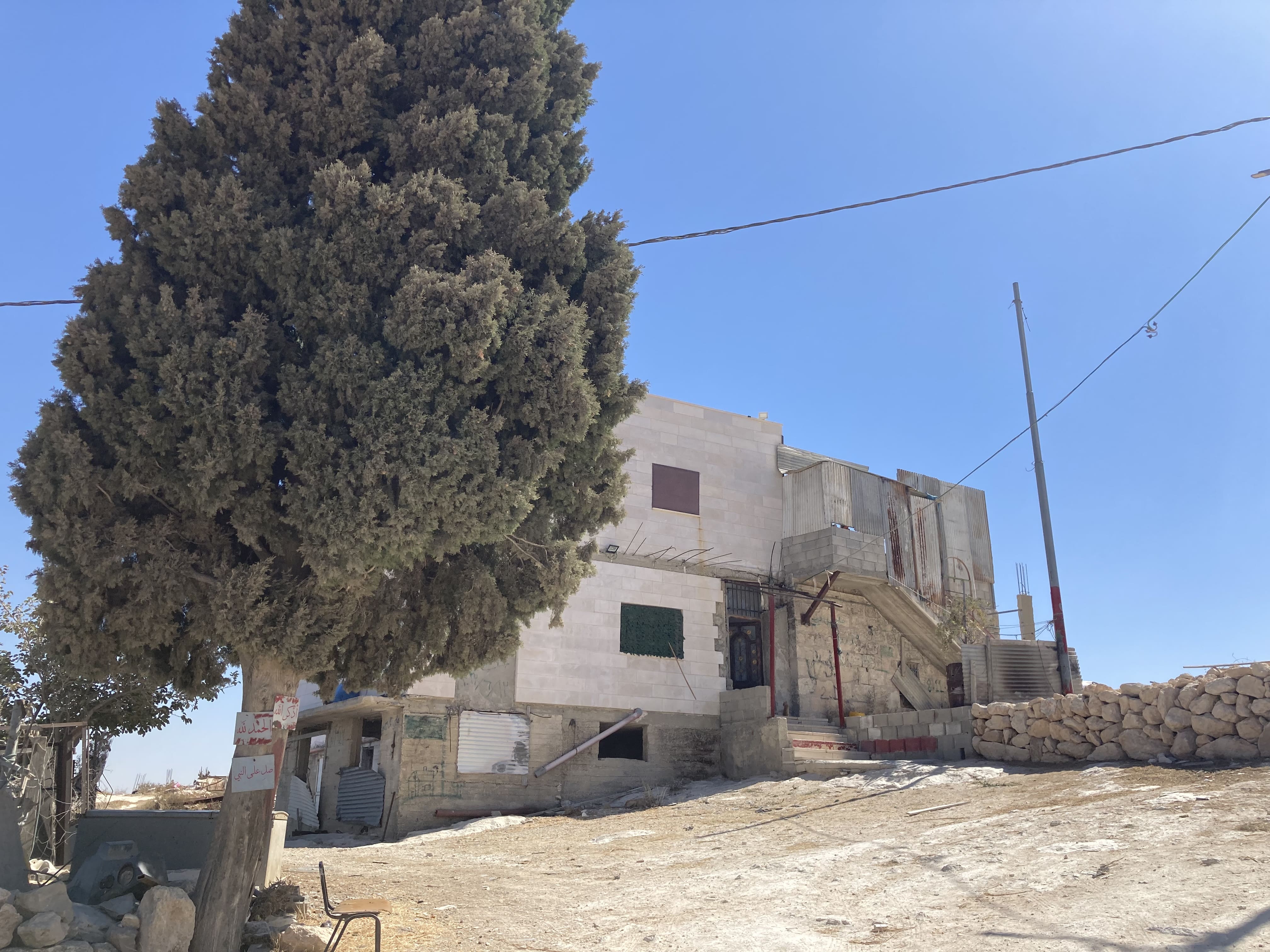 Yassin family home_EAPPI-min A concrete two story house with small square windows, without window panes. There is a tall pine tree in front and the sky is blue.