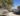 A photo of a dirt road with an olive tree hanging over it. In the back a small one story concrete house and some hills. The sky is blue.