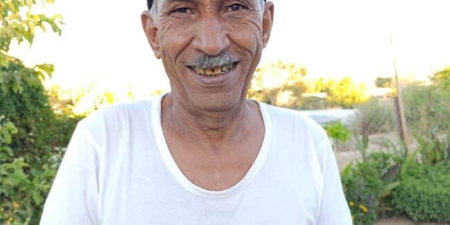 Man smiling wearing white t-shirt and black cap. His skin is tan and wrinkles are visible. Behind him a road and greenery.