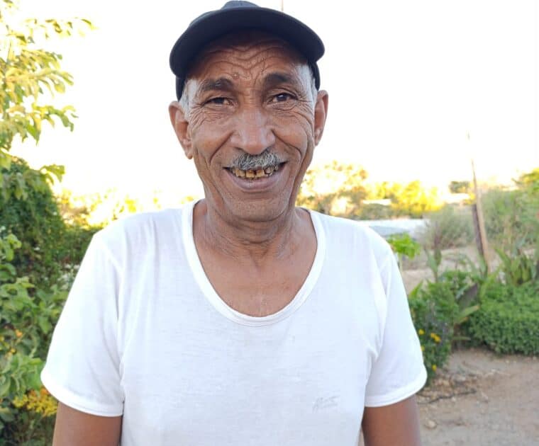 Man smiling wearing white t-shirt and black cap. His skin is tan and wrinkles are visible. Behind him a road and greenery.